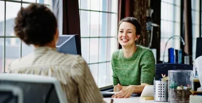 Two business women sitting at a desk talking.